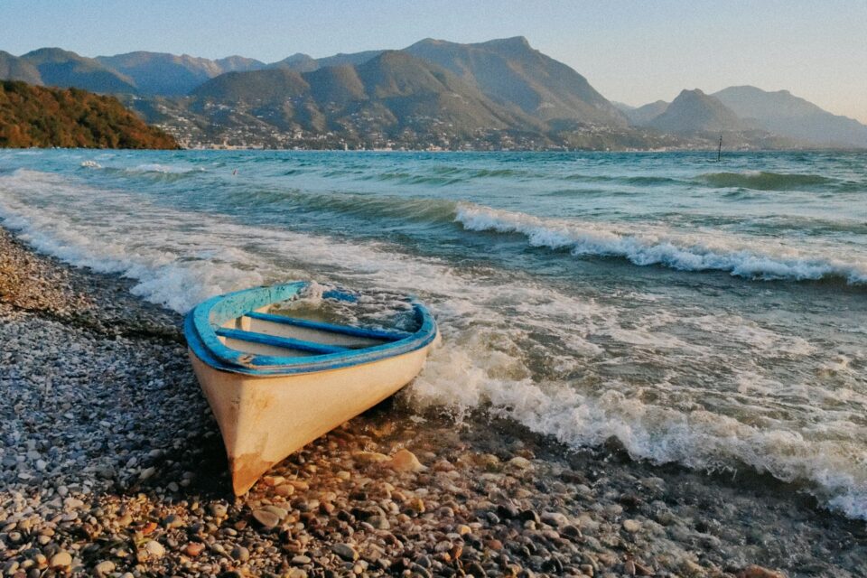 Piccola barca color crema e turchese sulla riva ghiaiosa, con onde del mare e montagne sullo sfondo.