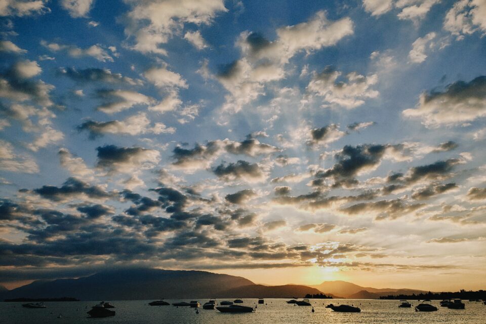 Tramonto sul mare con nuvole sparse, profilo di colline all’orizzonte e piccole barche ancorate in primo piano.