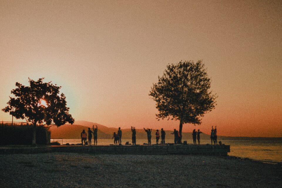 Persone in piedi su una passerella accanto a uno specchio d’acqua, con alberi e il sole basso dietro le colline.