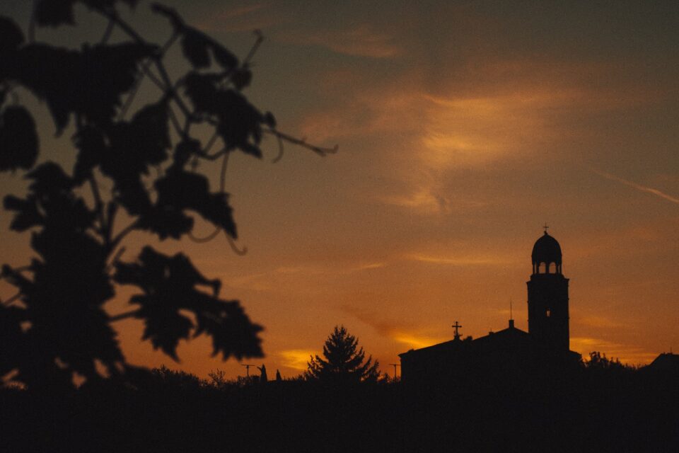 Sagoma scura di un edificio con torre campanaria e cupola, alberi in primo piano e cielo arancione al tramonto.