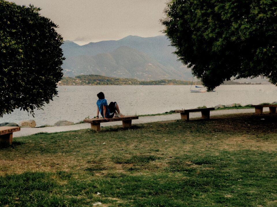 Persona seduta su una panchina in riva a un lago, con montagne e alberi sullo sfondo.