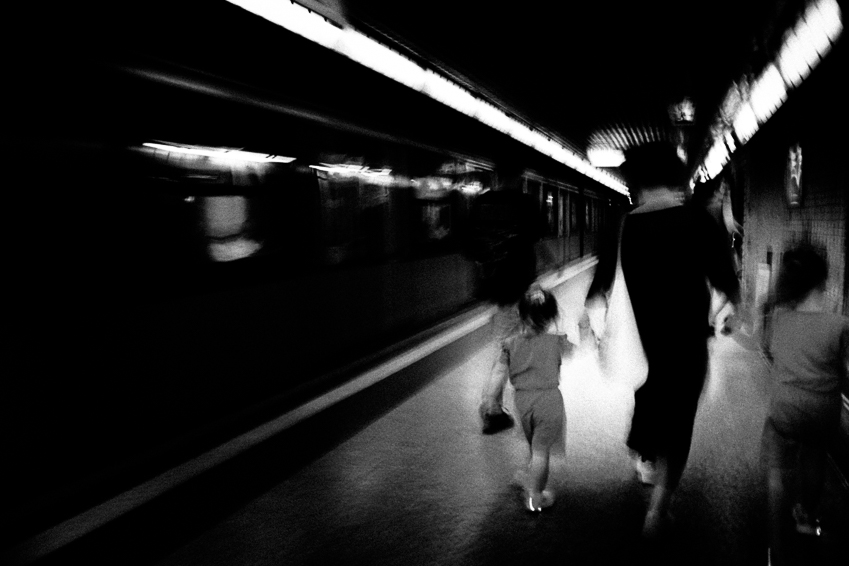 Persone camminano su una banchina della metropolitana accanto a un treno fermo, in un ambiente scuro illuminato da luci sul soffitto.