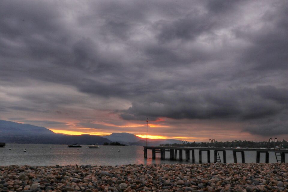 Spiaggia di ciottoli davanti a un lago, con un pontile, montagne e nuvole scure illuminate da una fascia di luce all'orizzonte.