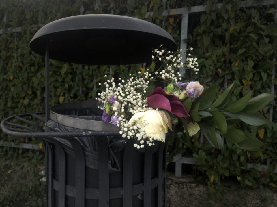 Cestino portarifiuti nero con coperchio, con un mazzo di fiori bianchi e colorati appoggiato sul bordo.