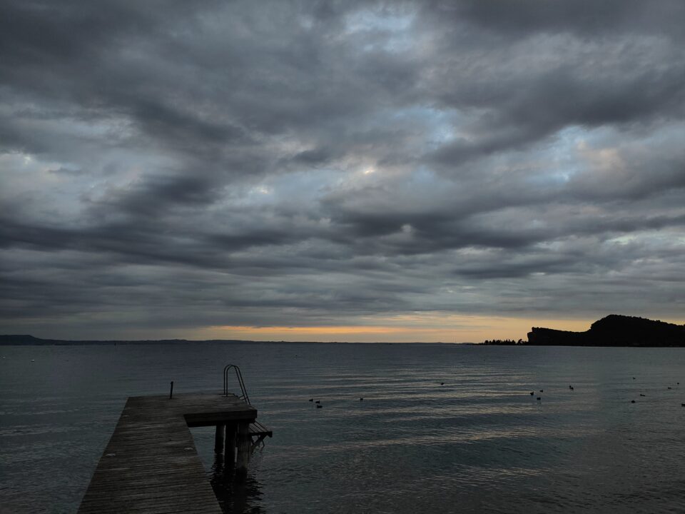 Pontile in legno con scaletta che si protende su un lago, sotto un cielo nuvoloso al tramonto.