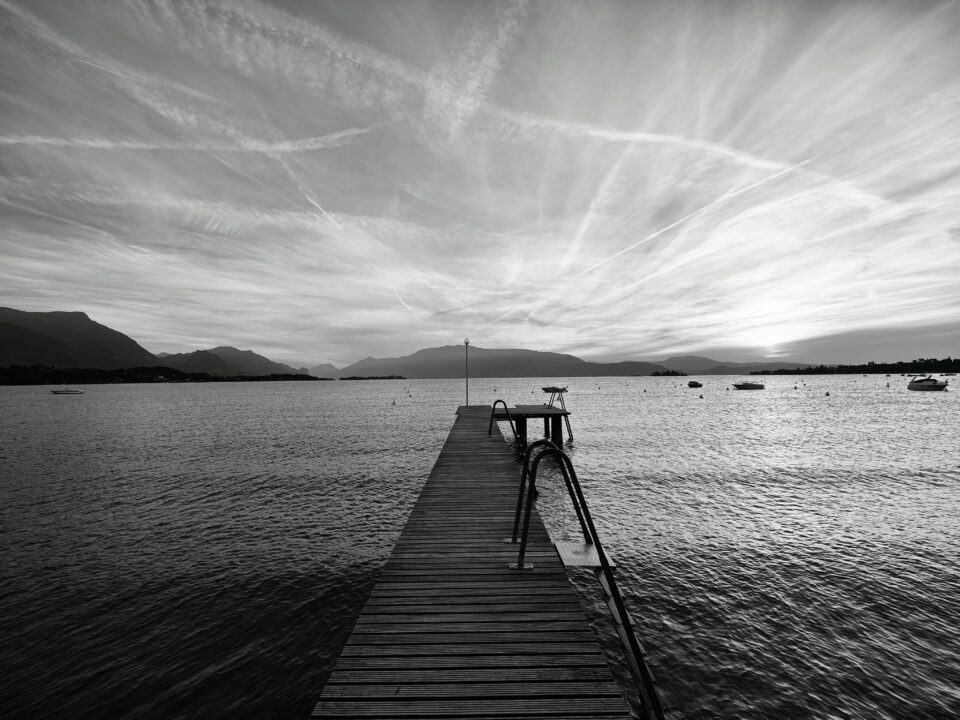 Pontile in legno che si estende su un lago, con montagne all’orizzonte, piccole barche sull’acqua e cielo nuvoloso.