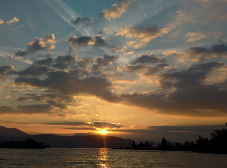 Tramonto sul lago, con il sole basso dietro le montagne, nuvole sparse nel cielo e riflessi dorati sull’acqua.