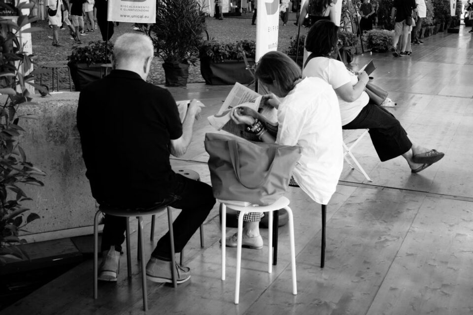 Tre persone siedono su sedie bianche; due consultano documenti a un tavolo, mentre una tiene un telefono. Altre persone e piante sullo sfondo.