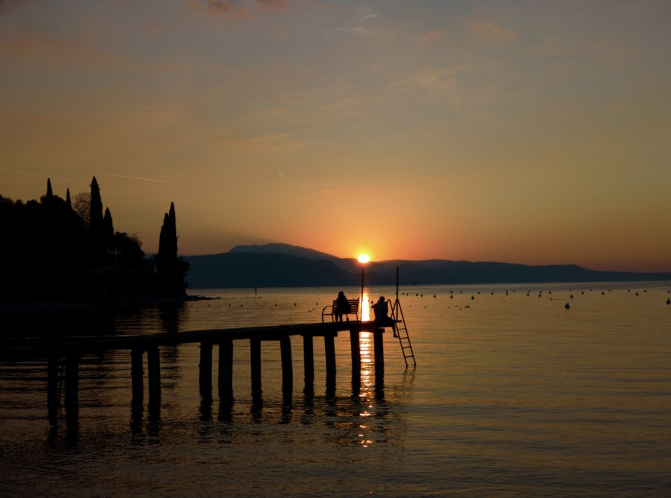 Tramonto sul lago con pontile in primo piano, persone sulla scaletta e sagome di alberi e montagne sullo sfondo.