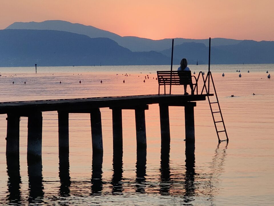 Persona seduta su una panchina in fondo a un pontile sul lago al tramonto, con una scaletta che scende nell’acqua e montagne sullo sfondo.