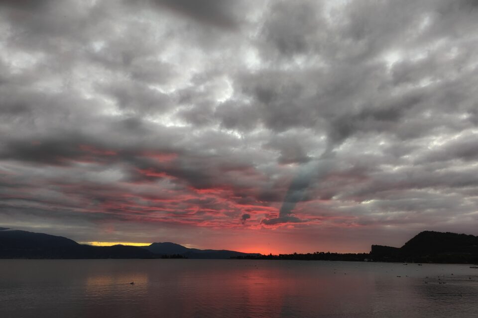 Lago calmo al tramonto, con montagne scure all’orizzonte e cielo coperto da nuvole grigie e rosse.