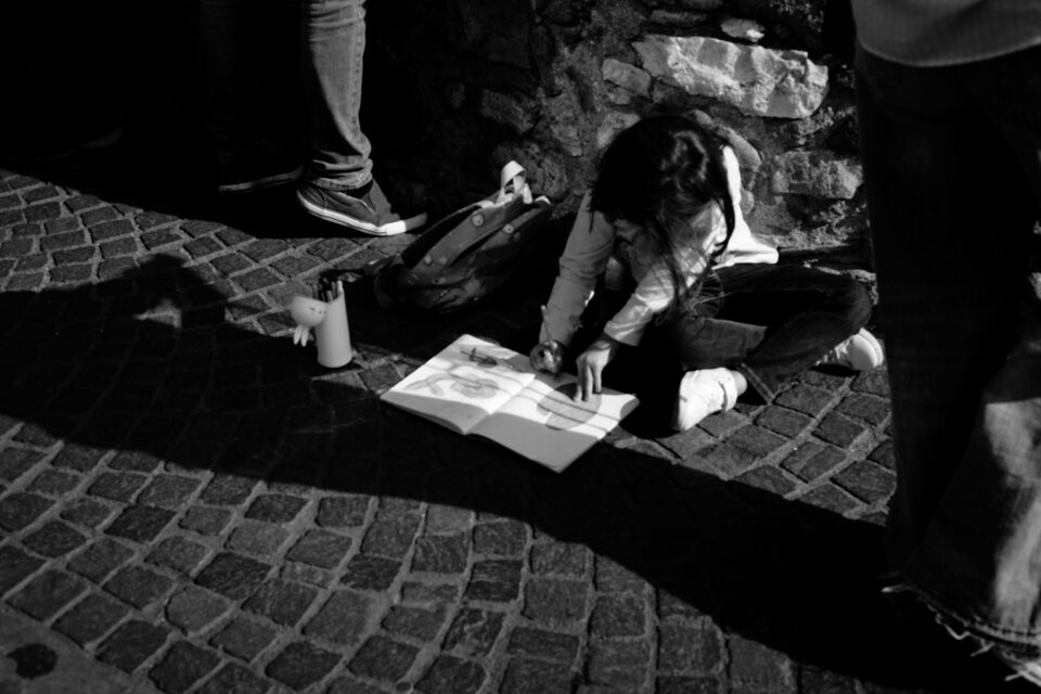 Bambina seduta su un selciato in pietra mentre disegna su un quaderno aperto. Accanto ci sono uno zaino e un portapenne, con gambe di persone in piedi attorno.