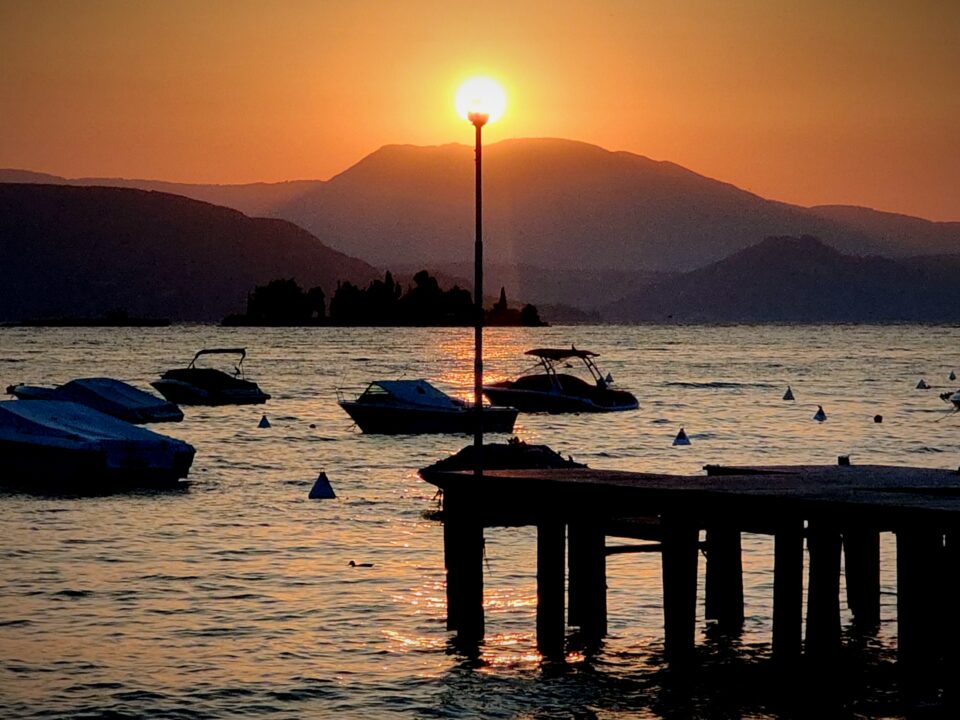 Tramonto su un lago circondato da montagne, con imbarcazioni ormeggiate, boe e un pontile in primo piano.