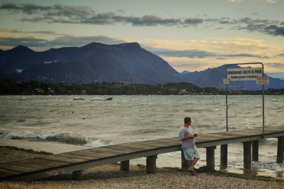 Una persona cammina su un pontile sopra uno specchio d’acqua mosso; sullo sfondo si vedono montagne, vegetazione e un cartello con testo.