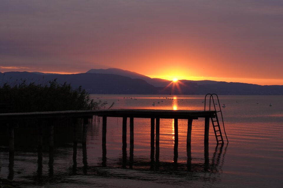 Tramonto su un lago con montagne sullo sfondo e un pontile in legno dotato di scaletta che si riflette nell’acqua.