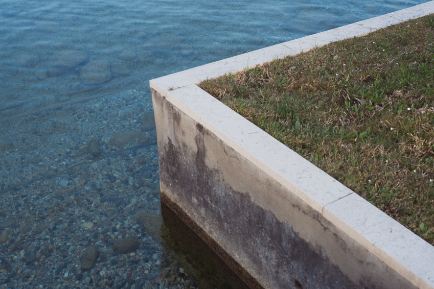 Bordo in cemento con erba sopra l’acqua limpida e ciottoli visibili sul fondo.