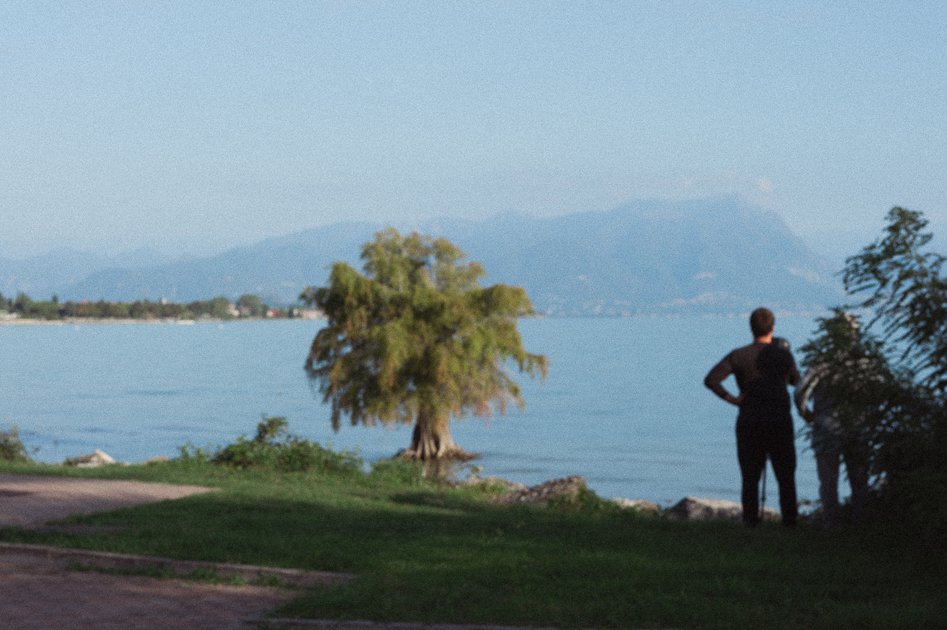 Persona in piedi sulla riva di un lago, con alberi in primo piano e montagne visibili sullo sfondo.