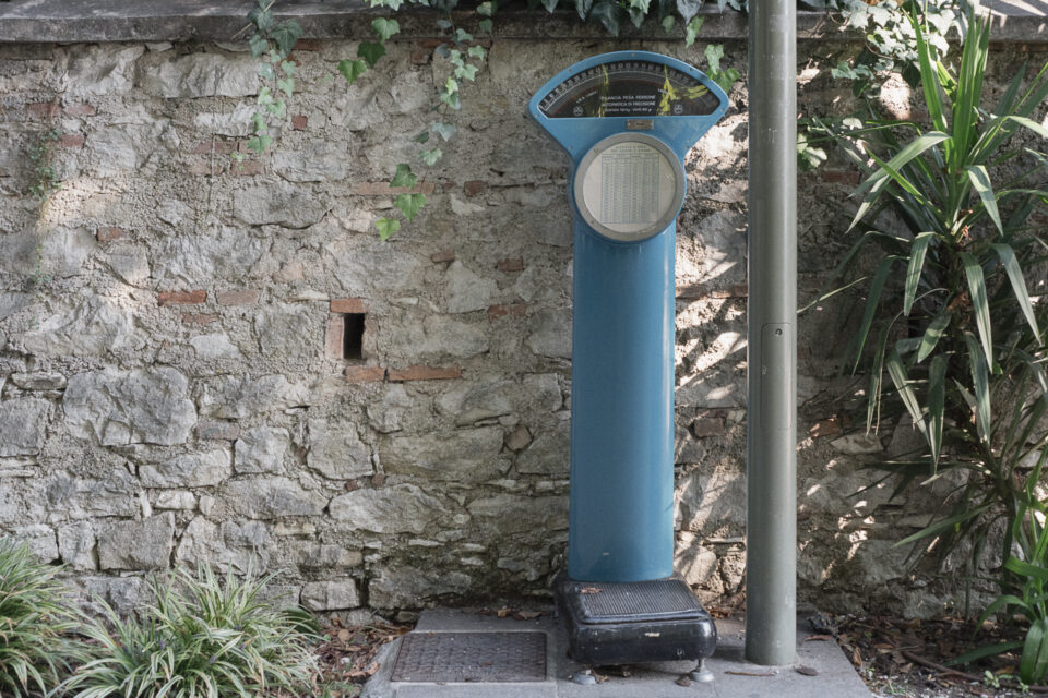 Bilancia pesapersone vintage a colonna blu con quadrante superiore, posizionata all'aperto davanti a un muro di pietra con piante.