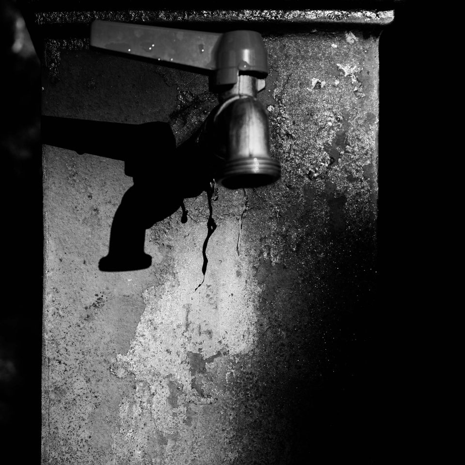 Rubinetto da esterno fissato su una parete scrostata, illuminato lateralmente mentre proietta una netta ombra scura.