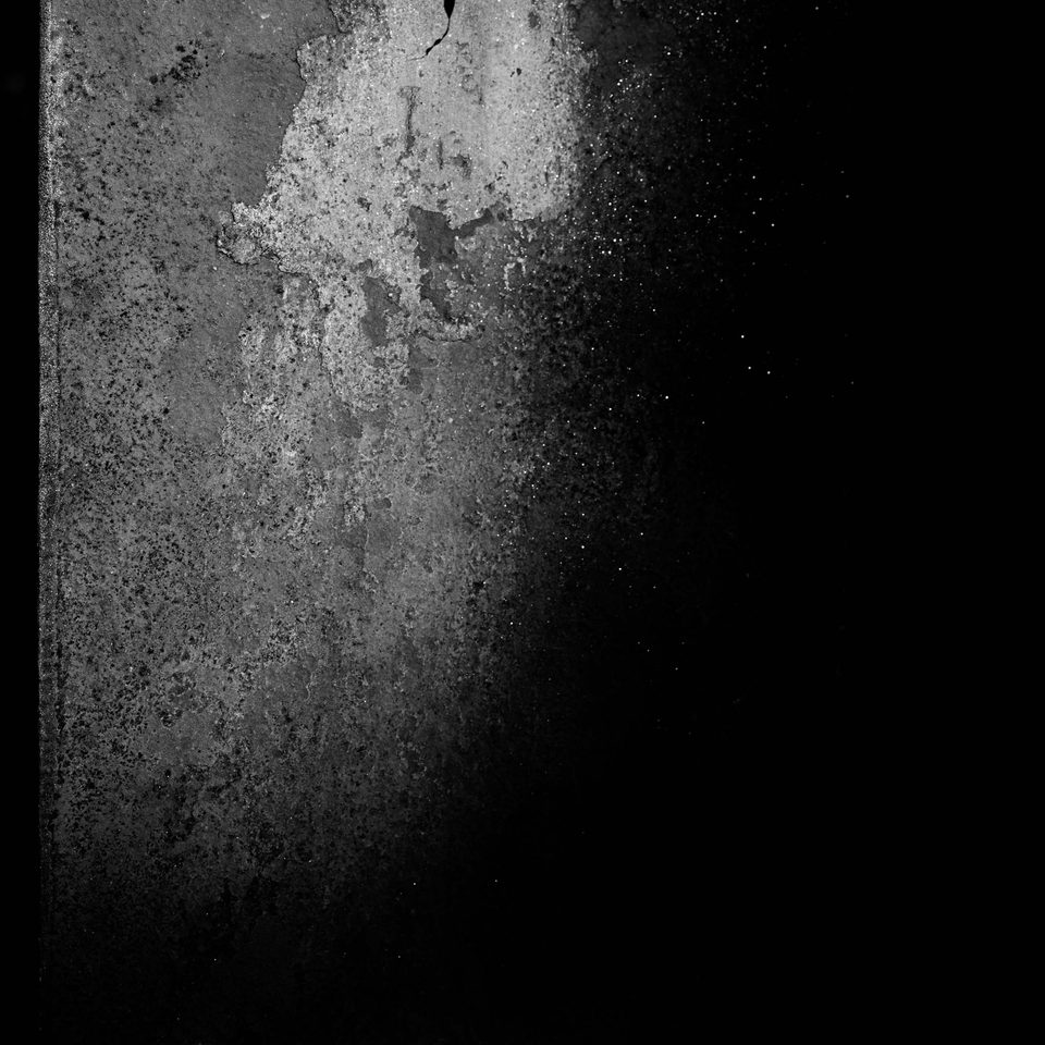 Superficie metallica o muraria ruvida e scrostata illuminata sul lato sinistro, che sfuma nel buio completo a destra.