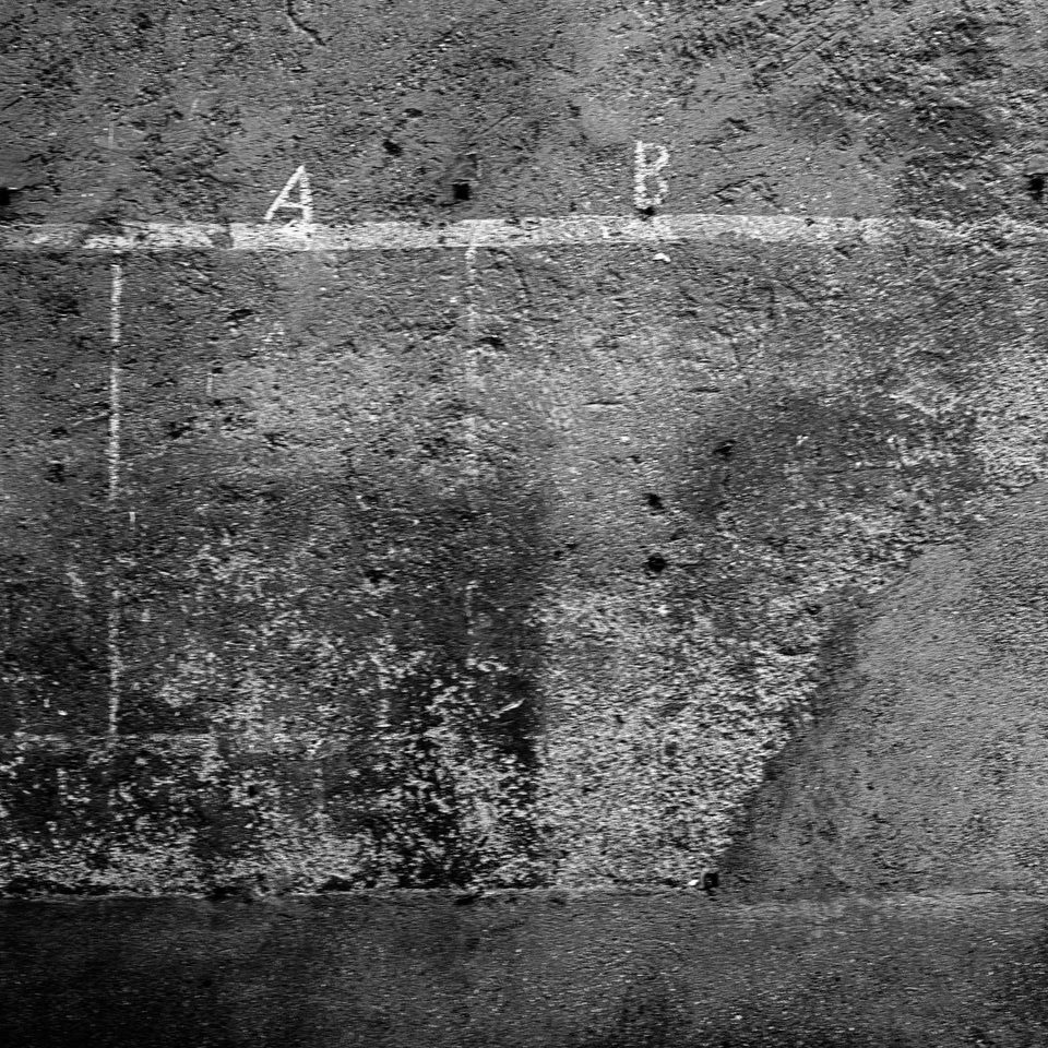 Muro in cemento ruvido e scrostato con una griglia tracciata a linee bianche, sormontata dalle lettere A e B.