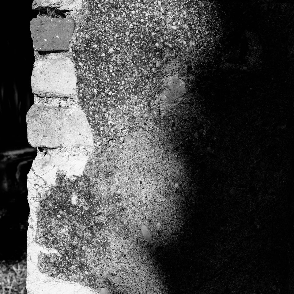 Primo piano in bianco e nero di una parete rovinata in cemento e mattoni parzialmente in ombra.