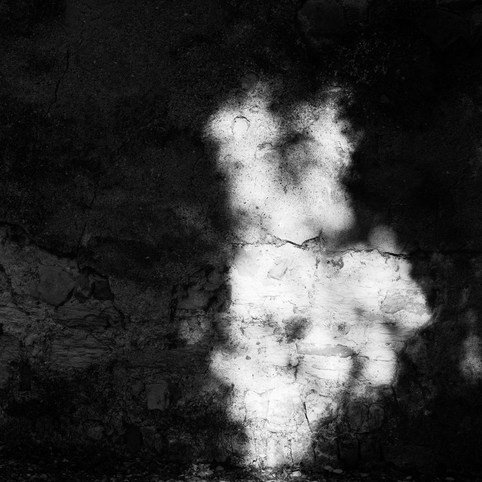 Muro in pietra e intonaco usurato in bianco e nero, avvolto nell'ombra con una macchia centrale illuminata dalla luce del sole.