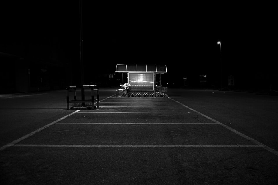 Area asfaltata vuota di notte, con una piccola struttura illuminata al centro, un lampione e una barriera in primo piano.